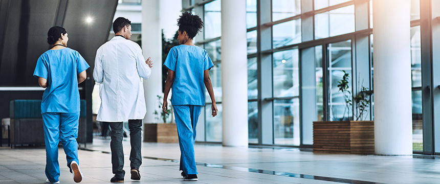 Three healthcare professionals, two in blue scrubs and one in a white coat, walk down a modern hospital corridor with large windows and indoor plants, resembling the sleek atmosphere of a corporate law office.
