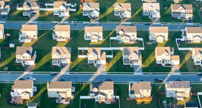Aerial view of a suburban neighborhood with rows of similar single-family houses, green lawns, and streets, casting long shadows in the sunlight—just miles away from a leading corporate law office.