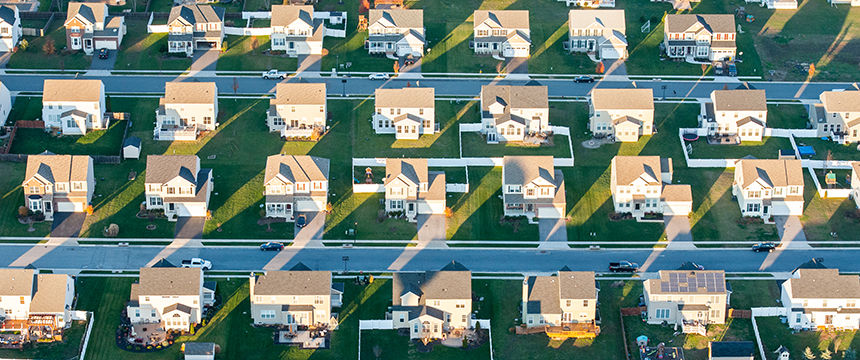 Aerial view of a suburban neighborhood with rows of similar single-family houses, green lawns, and streets, casting long shadows in the sunlight—just miles away from a leading corporate law office.