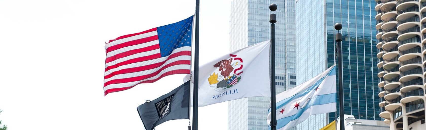 Four flags—including the U.S., Illinois state, POW/MIA, and Chicago city flags—fly on flagpoles in front of tall buildings housing law offices and lawyers in Chicago’s vibrant urban setting.