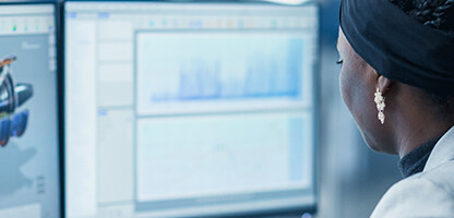 A woman sits at a desk in a law office, working on two monitors displaying technical graphics and charts—providing essential litigation support for intellectual property law cases.