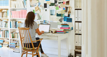 A woman sits at a desk working on a laptop in a home office, surrounded by bookshelves, binders, stacks of books, and a bulletin board—her workspace resembles the busy law offices of lawyers in Chicago.