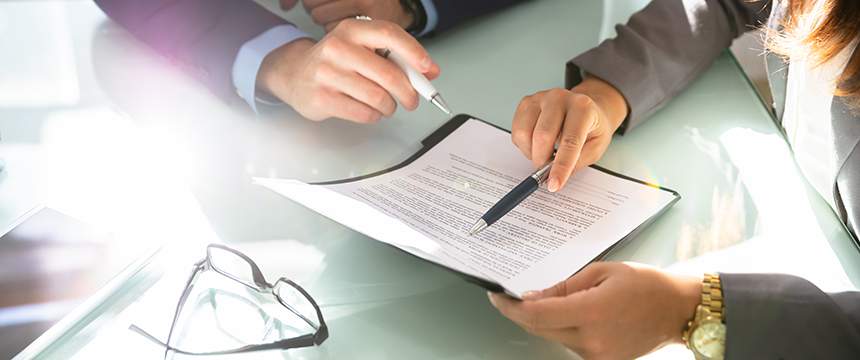 Two people in business attire, likely chicago lawyers, review and point at a document on a clipboard, with a pair of eyeglasses resting on the glass table nearby.