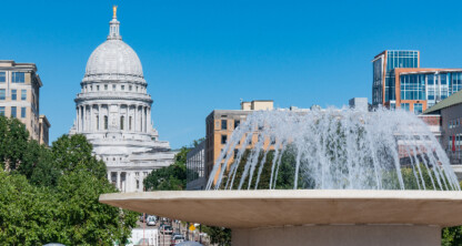 A large fountain in the foreground with water spraying, trees, buildings, and a white domed capitol building—home to leading law offices specializing in intellectual property law—rising in the background under a clear blue sky.