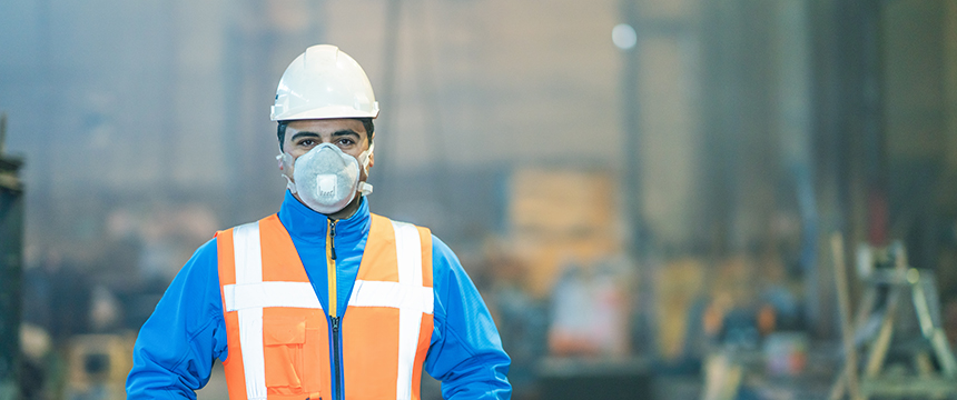 A construction worker wearing a white hard hat, orange safety vest, blue jacket, and a mask stands indoors at a work site—ideal imagery for chicago lawyers or law offices offering litigation support in construction cases.