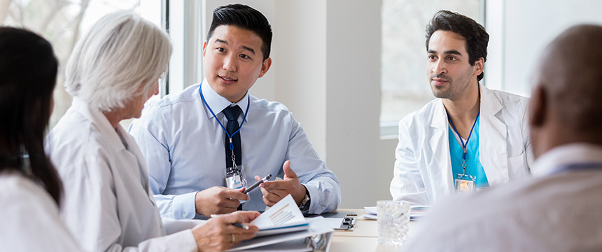 A group of doctors and a professional in a shirt and tie sit at a table, engaged in discussion with documents and notepads, resembling a strategic meeting often seen in law offices or among lawyers in Chicago.