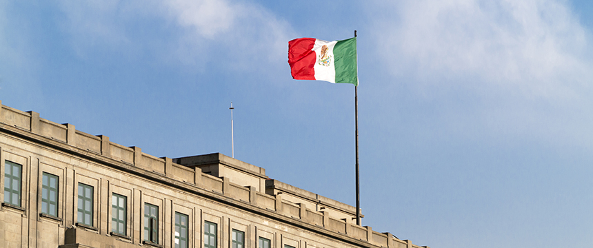 A Mexican flag flies atop a beige government building under a blue sky with some clouds, reminiscent of the professional presence found at top corporate law offices.