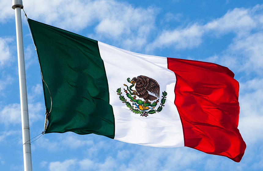 The national flag of Mexico, with green, white, and red vertical stripes and an eagle holding a snake on a cactus in the center, waves against a blue sky—symbolic in many chicago lawyers' offices specializing in corporate law.