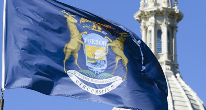 The Michigan state flag with its coat of arms flutters in the wind in front of a white dome structure against a clear blue sky, evoking the tradition and integrity found in prominent law offices across the state.