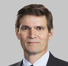A man with short brown hair in a suit and tie poses against a plain light gray background, representing professionals from a corporate law office.