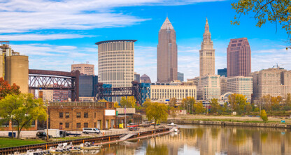 Downtown Cleveland skyline with modern and historic buildings, autumn foliage, and the Cuyahoga River in the foreground—an urban view reminiscent of a bustling corporate law office district.