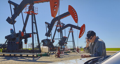 A man in a cap looks at papers and talks on the phone near several oil pumpjacks, possibly discussing litigation support, under a clear blue sky.
