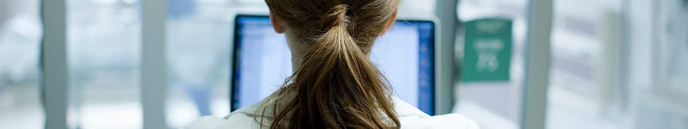 A person with brown hair tied in a ponytail sits facing a computer monitor in a bright indoor setting, typical of modern law offices.
