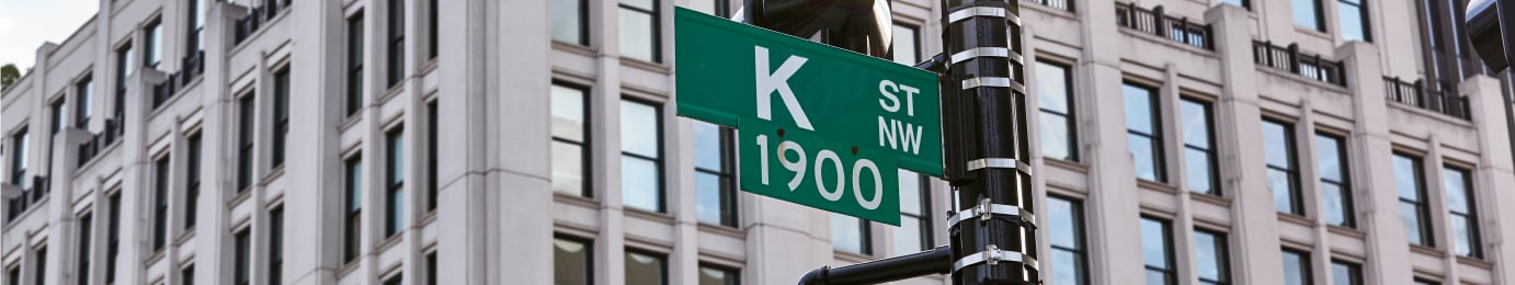 Green street sign reads "K St NW 1900" against a backdrop of a modern office building with large windows, evoking the professionalism of a corporate law office.