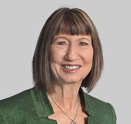 A woman with straight, shoulder-length brown hair and bangs, wearing a green jacket and a necklace, smiles at the camera against a plain gray background, embodying the professionalism found in top chicago lawyers.