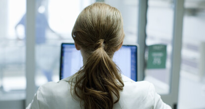 A person with brown hair in a ponytail, seen from behind, sits at a desk in front of a computer monitor in a professional office, perhaps working among Chicago lawyers or researching intellectual property law.