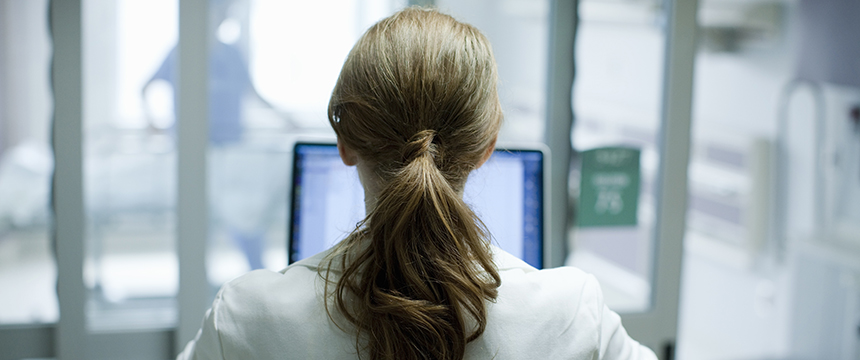 A person with brown hair in a ponytail, seen from behind, sits at a desk in front of a computer monitor in a professional office, perhaps working among Chicago lawyers or researching intellectual property law.