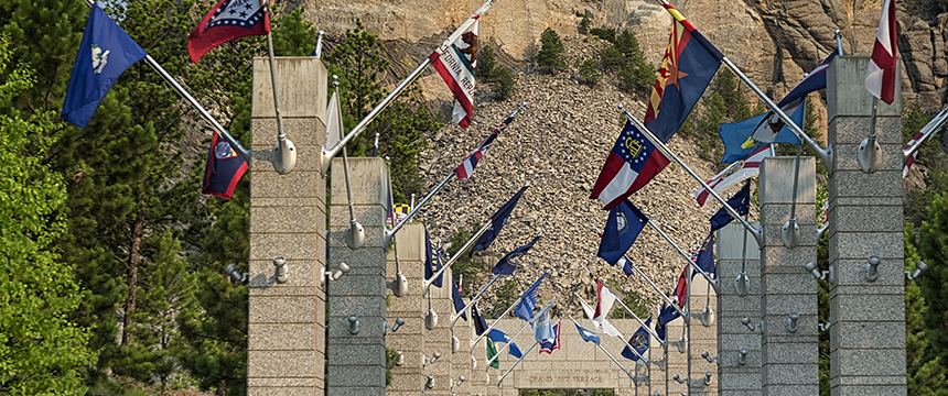 Rows of granite pillars displaying various state flags line a walkway leading toward a rocky hill surrounded by trees, evoking the prestige and tradition often found at a distinguished corporate law office.