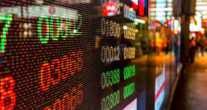 Brightly lit digital stock ticker displays numbers in red and green, reflected on a glass wall near bustling law offices in a busy urban street setting.