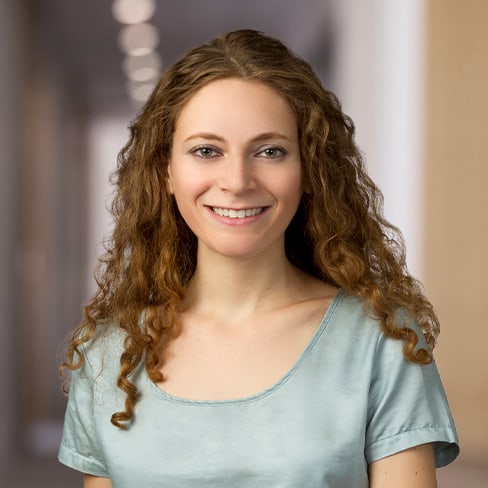 A woman with long curly brown hair, wearing a light blue short-sleeved top, smiles while standing in a blurred hallway of a corporate law office.
