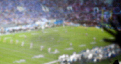 A blurry image of a football game in progress at a stadium, with players on the field and spectators in the stands, shot near a corporate law office bustling with activity.