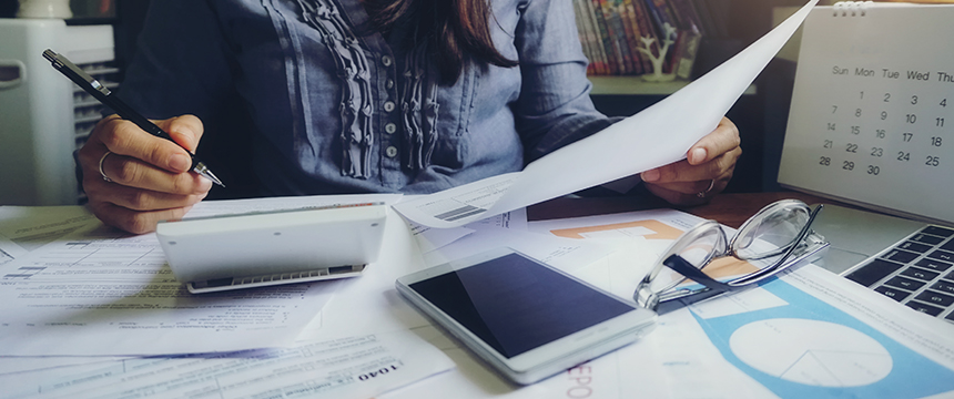 A person sits at a cluttered desk reviewing documents, using a calculator, with a smartphone, eyeglasses, and a calendar nearby—typical of busy law offices specializing in intellectual property law.