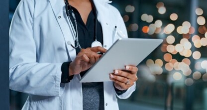 A doctor in a white coat and stethoscope uses a tablet device, the blurred lights hinting at a corporate law office collaborating on healthcare innovation.