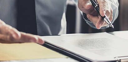 Two people sit at a desk, one holding a pen and gesturing toward a clipboard with documents, suggesting a discussion or signing of paperwork in a corporate law office.