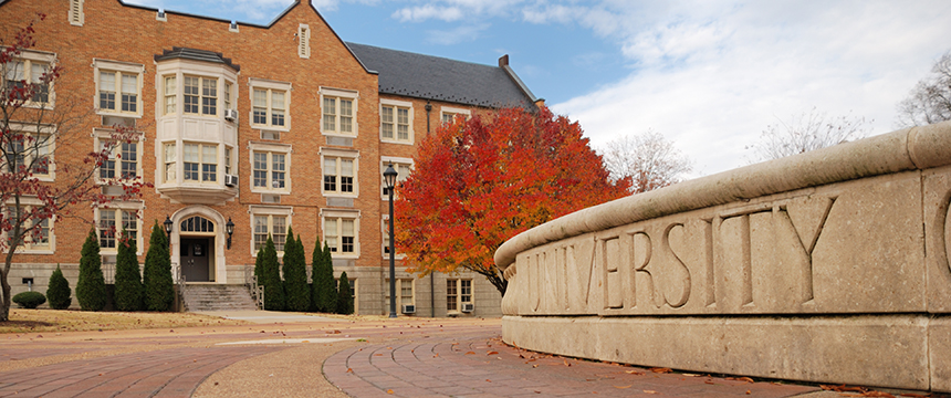 A brick university building with white trim stands behind a stone sign engraved with "UNIVERSITY" and a tree with red autumn foliage, offering an inspiring backdrop often frequented by Chicago lawyers and law offices for litigation support events.