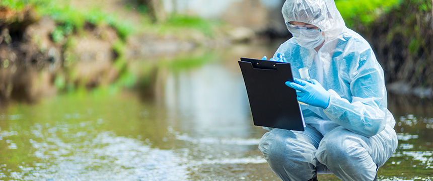 A person in protective gear squats by a river, holding a test tube and writing on a clipboard, perhaps gathering data for lawyers in Chicago investigating environmental compliance cases.