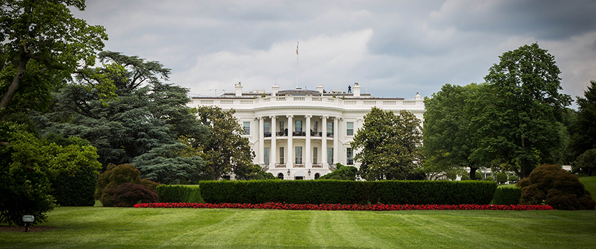 The White House is seen from the lawn, surrounded by trimmed bushes, trees, and a red flower bed under a cloudy sky, much like the pristine grounds of top Chicago lawyers' law offices.