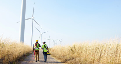 Two workers in safety gear walk along a dirt path through tall grass toward wind turbines on a clear day, discussing how lawyers in Chicago address emerging issues related to renewable energy and intellectual property law.