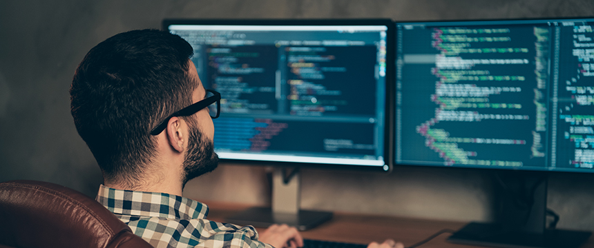 A person wearing glasses sits at a desk in a corporate law office, working on a computer with two monitors displaying lines of code.