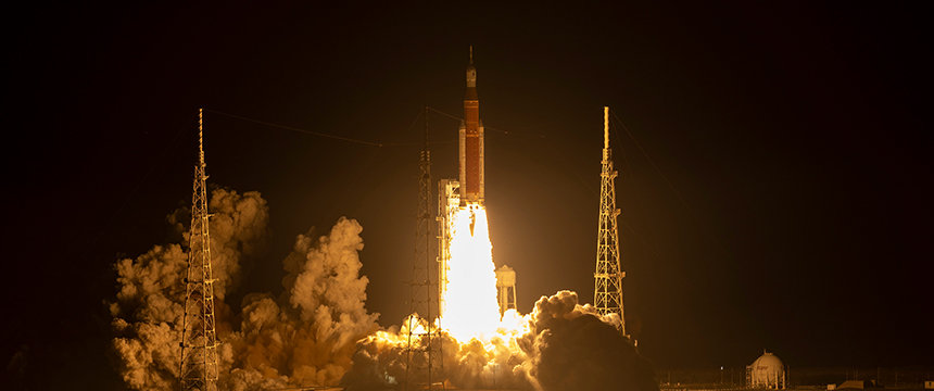 A rocket launches from a pad at night, with bright flames and smoke billowing beneath it, surrounded by support towers—much like law offices providing litigation support for complex intellectual property law cases.