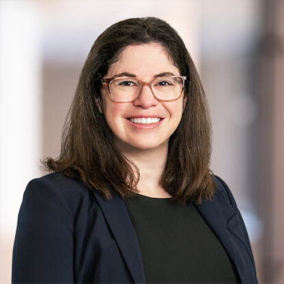 Woman with long brown hair and glasses, wearing a dark blazer and black top, smiling in front of a blurred corporate law office background.