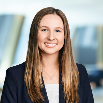 A woman with straight, light brown hair wearing a navy blazer and white top smiles at the camera against a blurred corporate law office background.