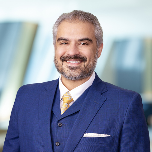 A man with gray hair and beard, wearing a blue suit, white shirt, and yellow tie, smiles in front of a blurred corporate law office background.