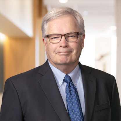 An older man with gray hair and glasses wearing a dark suit, blue patterned tie, and light blue shirt stands in a well-lit law office hallway, representing experienced Chicago lawyers.