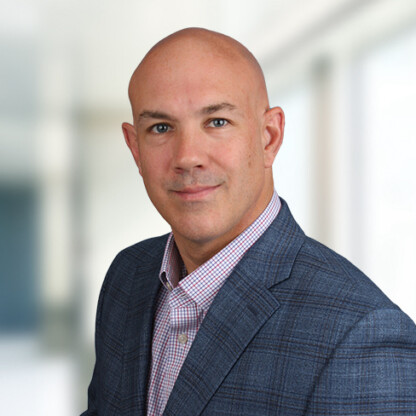 A bald man in a checked shirt and blue blazer poses for a professional headshot against a blurred law offices background.