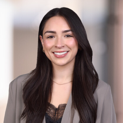 Woman with long dark hair wearing a taupe blazer and black lace top, smiling in a softly blurred law offices setting.