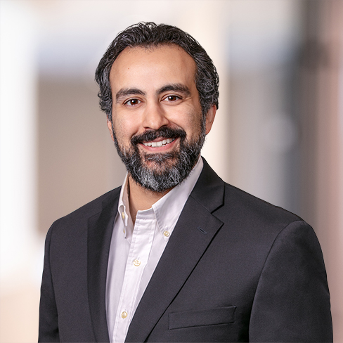 A man with dark hair and a beard wearing a dark suit jacket and white shirt smiles at the camera against a blurred indoor background, reflecting the professional atmosphere of a corporate law office.