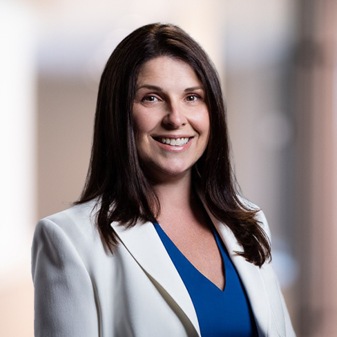 A woman with long brown hair, wearing a white blazer over a blue top, smiles at the camera against a blurred corporate law office background.