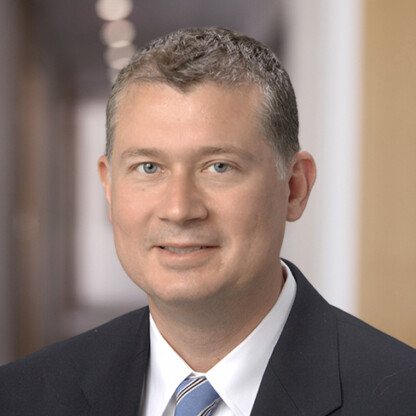 A man with short, graying hair wearing a dark suit, white shirt, and striped tie stands posed in the blurred hallway of a corporate law office.