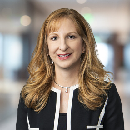 A woman with long, wavy blonde hair wearing a black jacket with white trim and a silver necklace, posing confidently in a corporate law office setting.
