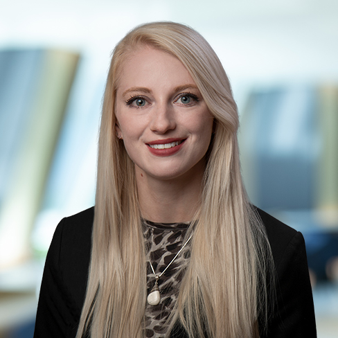 A woman with long blonde hair wearing a black blazer and patterned top, smiling at the camera in a blurred corporate law office—perfectly capturing the professional spirit of Chicago lawyers.