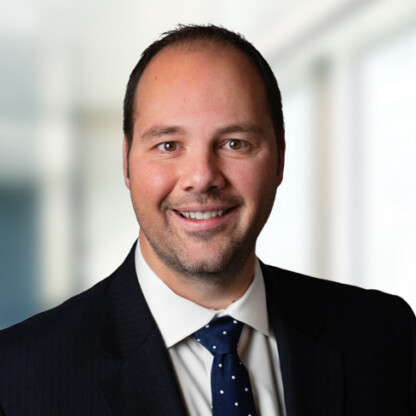 A man in a dark suit and polka dot tie smiles at the camera, standing in front of a blurred corporate law office background.