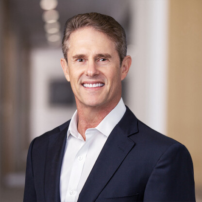 A man in a dark suit jacket and white shirt smiles while standing in a brightly lit, blurred corporate law office hallway.