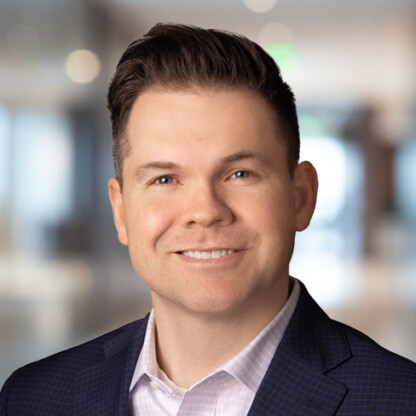 A man in a dark suit jacket and light shirt smiles at the camera, with a blurred corporate law office in the background.