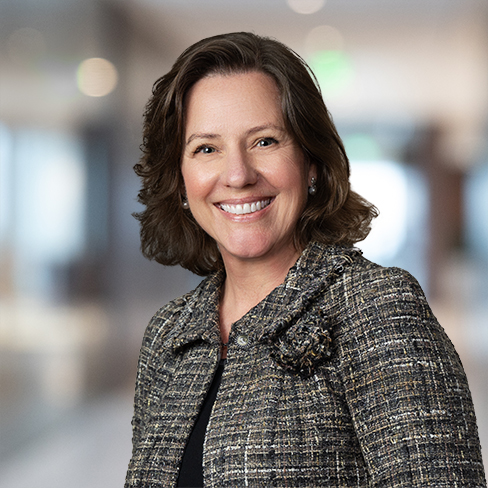 Woman with shoulder-length brown hair wearing a textured blazer, smiling, standing in a blurred corporate law office environment.