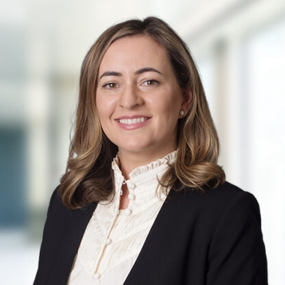 A woman with shoulder-length brown hair, wearing a black blazer and white ruffled blouse, smiles at the camera in a bright, blurred indoor setting typical of modern law offices.